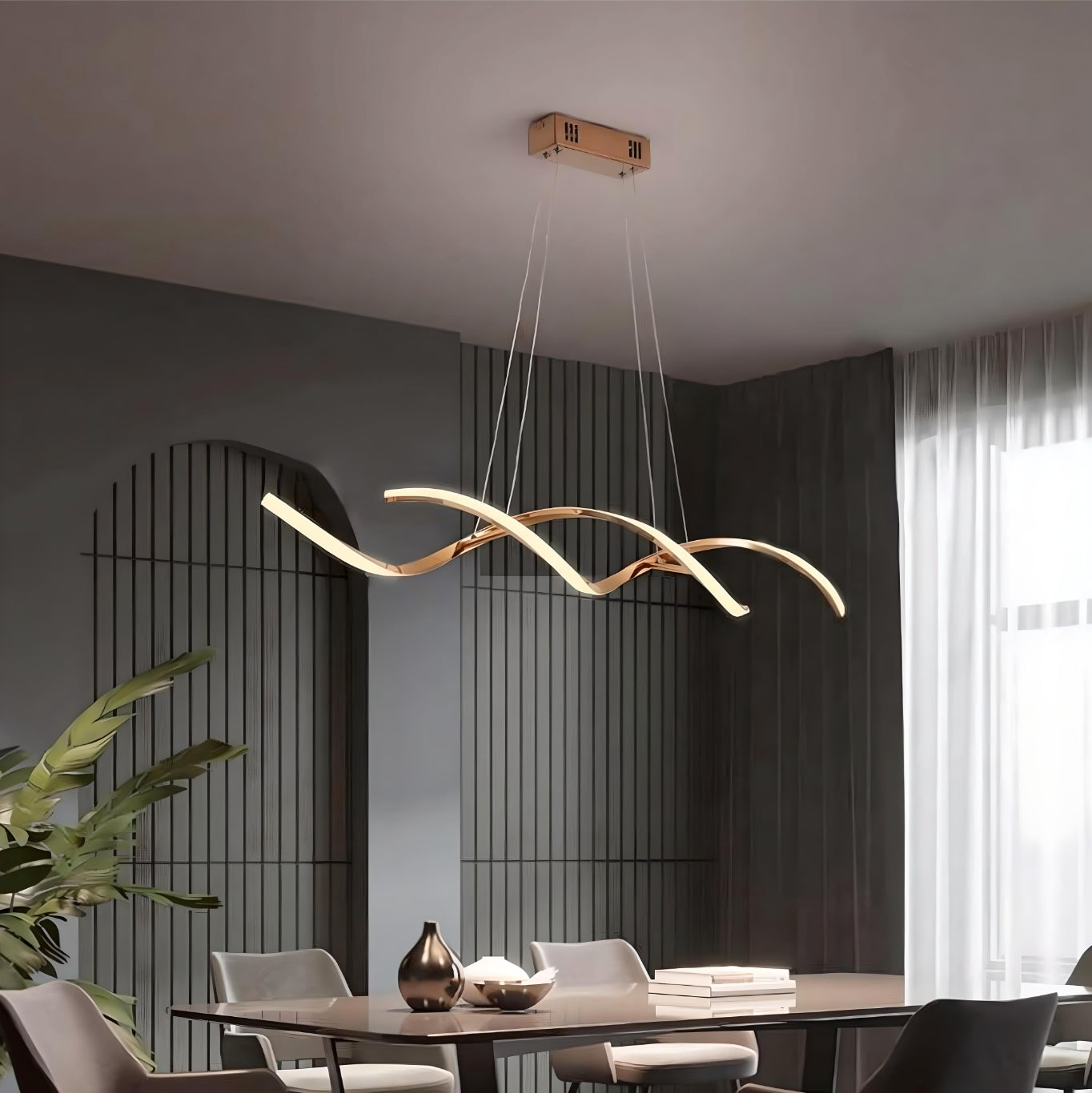 Suspension design LED dorée en forme de vagues entrelacées, suspendue au plafond d’un intérieur moderne avec murs gris et grande fenêtre lumineuse.
